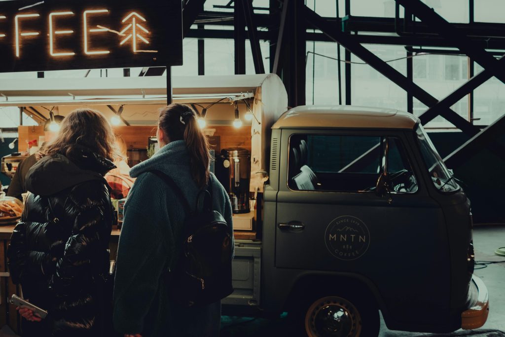 Back view of unrecognizable friends near coffee van under shiny neon signboard in evening